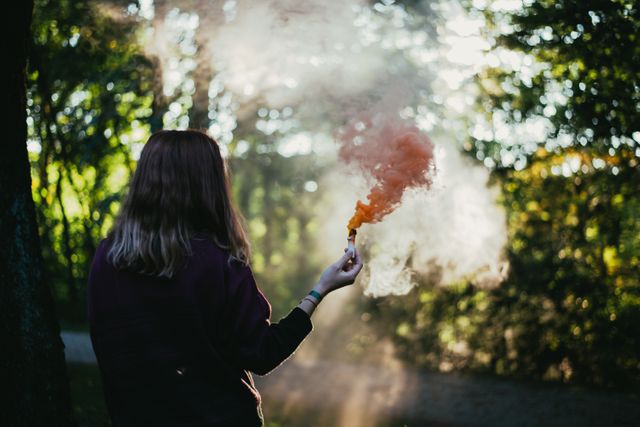Person holding an orange smoke bomb in a forest, surrounded by trees and fog. The lighting creates a serene and mystical atmosphere. Perfect for themes related to adventure, outdoor activities, forest exploration, nature, and tranquility.