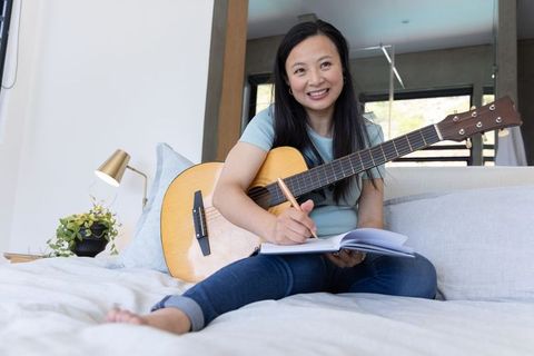 Asian woman songwriting on bed with acoustic guitar and notebook smiling in cozy bedroom