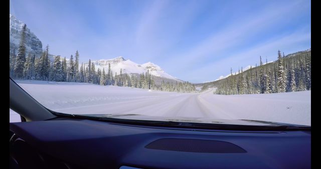 View from inside a vehicle showing a snow-covered road surrounded by evergreen trees and mountains in the distance. The perspective suggests a winter road trip through a serene, snowy landscape.