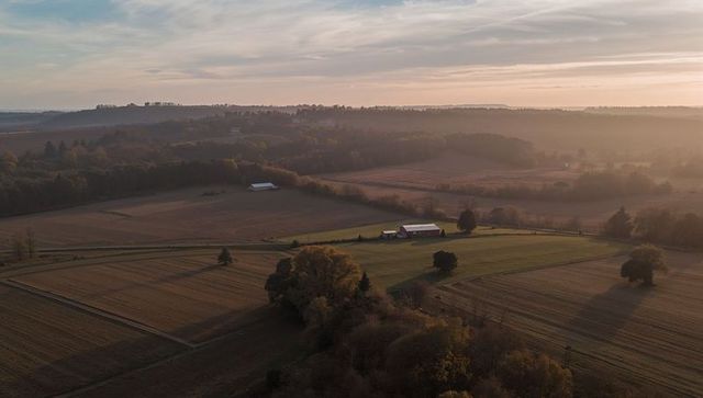 Golden-hour aerial capturing red-roof barn and fenced pasture on rolling countryside farmland