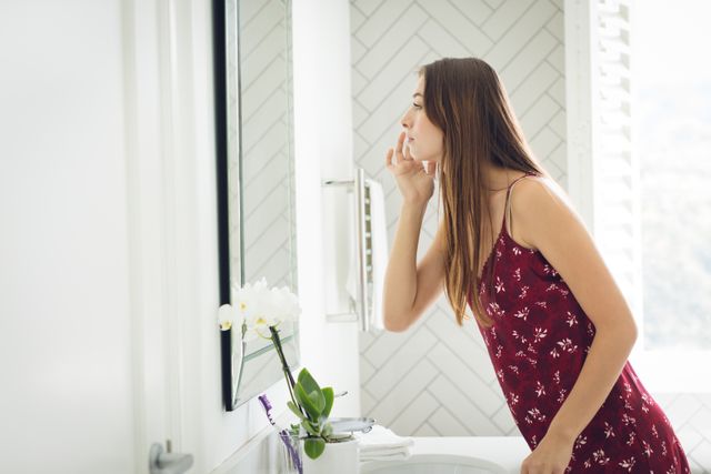 Young woman in a floral dress examining her face in the bathroom mirror. The bathroom features a white, modern design with a potted orchid on the counter. Ideal for use in articles or advertisements related to skincare, beauty routines, self-care, and home lifestyle.