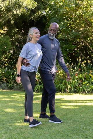 Senior couple walking arm-in-arm across sunlit garden lawn, outdoor fitness and wellbeing