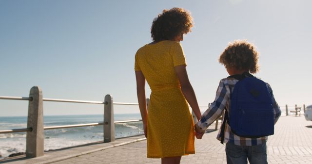 Happy biracial mother and son holding hands walking on sunny promenade by the sea, copy space. Motherhood, childhood, togetherness, summer, vacations and free time, unaltered.