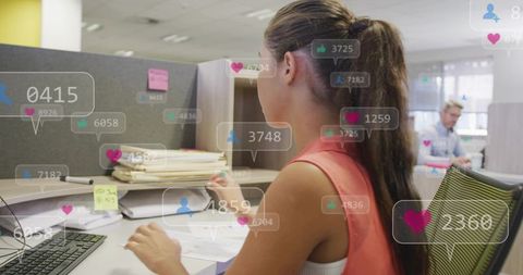 Young woman sorting papers and typing at cubicle with social media notification overlays