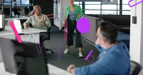 Walking woman in green blazer pausing midstep in modern open-plan office with coworkers
