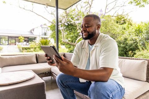 African man smiling while using tablet on outdoor rattan sofa at modern home terrace