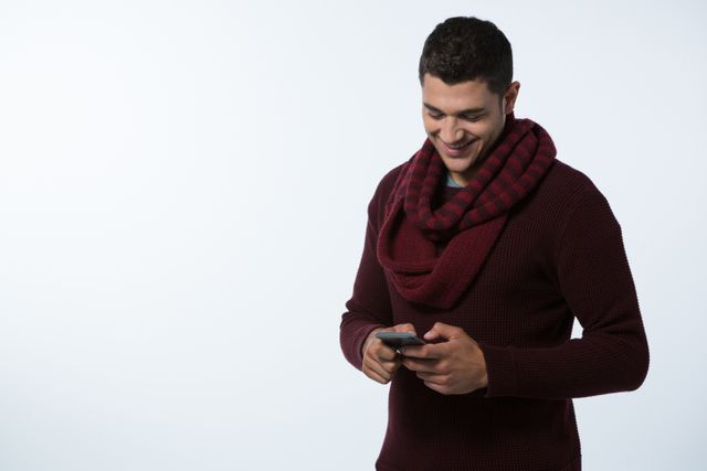 This image depicts a young man dressed in winter clothing, including a scarf and sweater, smiling while using a mobile phone. The white background makes it ideal for use in advertisements, websites, or social media posts related to technology, communication, fashion, or lifestyle. It can also be used in promotional materials for mobile apps or winter clothing brands.