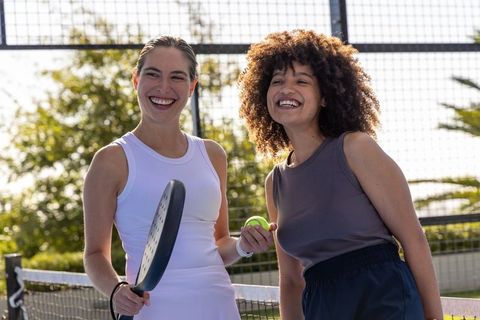 Smiling diverse women playing paddle tennis on outdoor court holding racket and ball