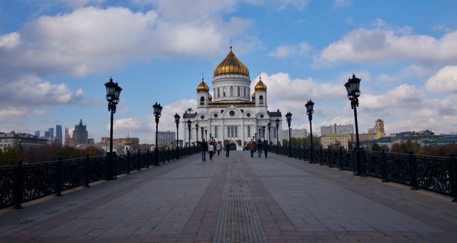 Tourists walking towards Cathedral of Christ the Saviour in Moscow via pedestrian bridge. Golden domes and detailed facade stand out under partly cloudy sky. Ideal for use in travel blogs, articles on Russian architecture, history documentaries, or promotional travel content.