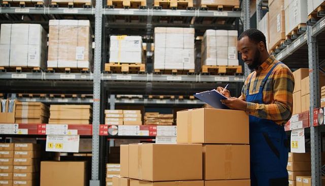 Warehouse worker checking inventory on clipboard amid stacked boxes and industrial shelving