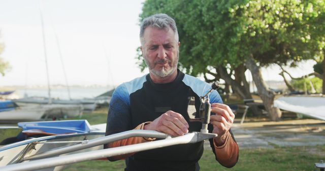 An elderly man is performing maintenance on a sailboat outdoors in a marina. This is ideal for conveying themes of leisure activities, boating, outdoor hobbies, nautical enthusiasm, and the active lifestyle of mature adults. Useful for articles and advertisements for boating equipment, senior activity engagement, and marine maintenance tips.