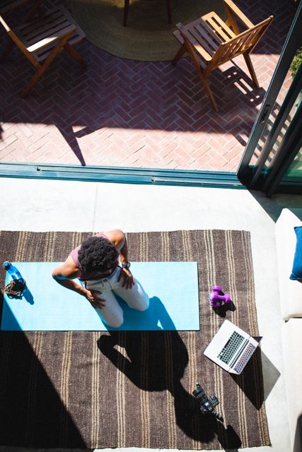 This image shows a high angle view of an African American man exercising at home on a yoga mat. He is surrounded by a laptop, a camera, and a water bottle, suggesting he is vlogging his workout routine. This image can be used for content related to home fitness, health and wellness, influencer marketing, and technology in fitness.