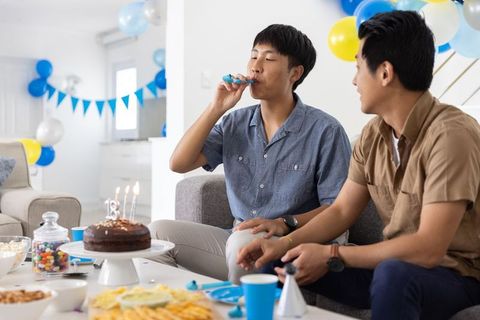 Asian men celebrating birthday at home blowing party horn with chocolate cake and balloons