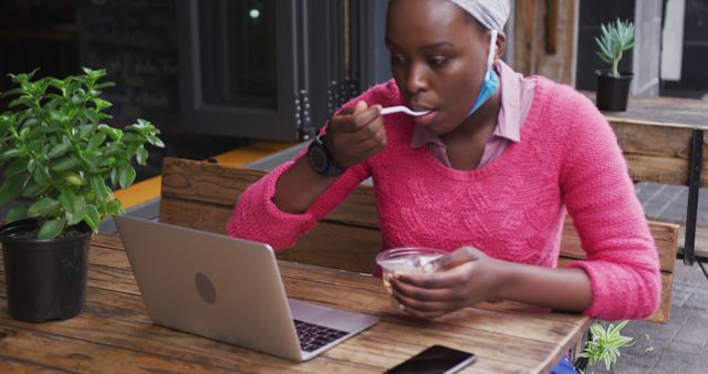 Young woman eating a snack and working on a laptop in a cafeteria. She is wearing a pink sweater and sitting by a table with plants. Ideal for images relating to multitasking, modern work habits, remote work, urban lifestyle, productivity, and technology in everyday life.