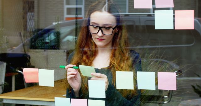 Young woman stands by a window, attaching and arranging sticky notes to organize tasks in an office setting. This image can be used for depicting team meetings, project planning sessions, corporate strategy discussions, or illustrating organizational tools. Perfect for business-related projects, articles on workflow optimization, or any scenario requiring imagery about creative thinking and meticulous planning.