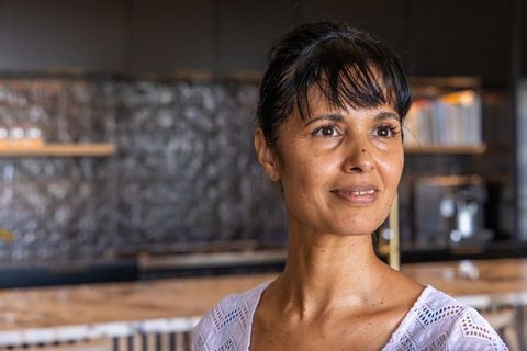 Mature african woman smiling and standing at modern café wooden counter wearing white top