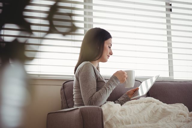 Woman sitting on couch, wrapped in blanket, using tablet and drinking coffee. Ideal for lifestyle blogs, technology use, home comfort, and relaxation themes.