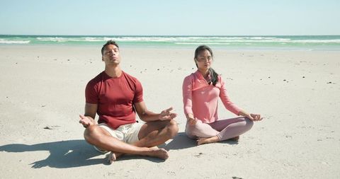 Diverse couple meditating cross-legged on sunny beach, practicing seaside mindfulness