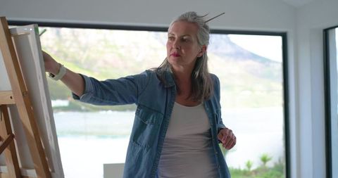 Contemporary woman painting at easel with lake view through large window in sunlit studio