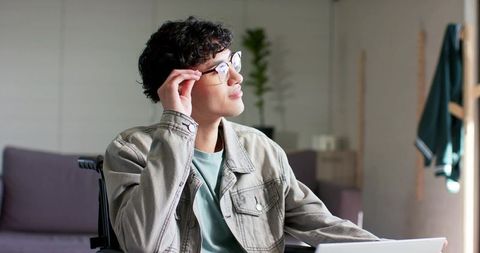 Young man in wheelchair adjusting glasses while working on laptop in bright home office