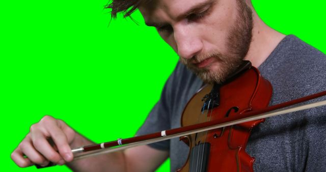 Young man focusing while playing violin against green screen background. Could be used for music education, promotional material for music lessons, or advertising musical instruments. Perfect for visual effects, allowing for easy background removal or replacement.