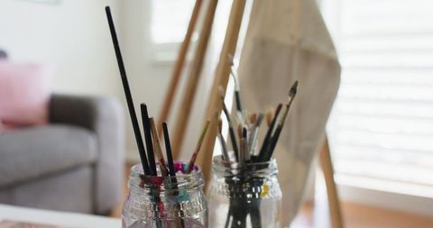 Paintbrushes in Glass Jars on Sunlit Art Table with Easel and Window Blinds
