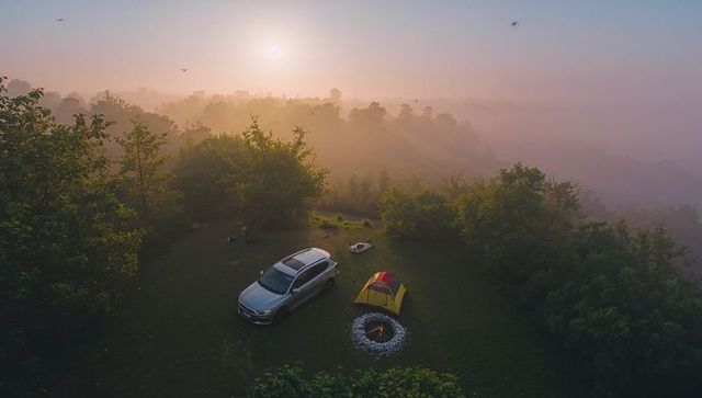 Sunrise hilltop camping with suv and yellow-red tent beside glowing campfire over misty ridge