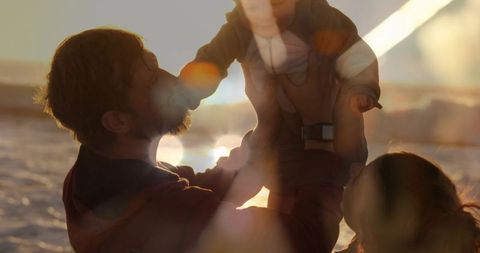 Father lifting baby into sunset on beach during golden hour, family warmth and tenderness