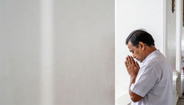 Middle-aged man praying and bowing head at window ledge with folded hands for quiet reflection