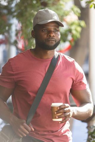 African american man walking urban sidewalk holding coffee cup and satchel in sunlight