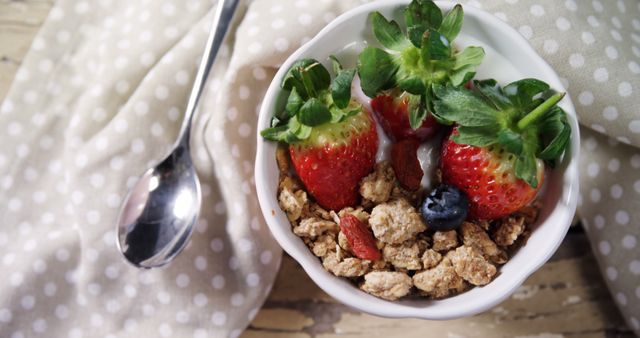 Nutritious breakfast bowl with fresh strawberries, blueberries, and granola on a polka dot cloth, accompanied by a spoon. Ideal for illustrating healthy eating habits, diet meal plans, breakfast menus, and food blogs focusing on nutritious breakfast options.
