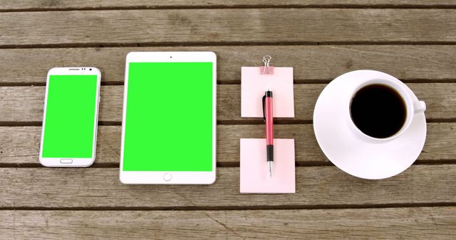 Top view of smartphone, tablet, pen, and cup of coffee arranged neatly on wooden desk. Green screens on smartphone and tablet for easy image insertion. Suitable for showcasing digital devices, office setups, or inclusive workspaces.