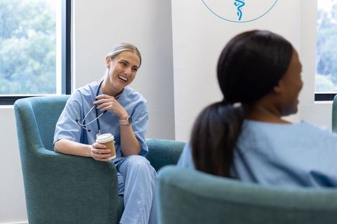 Nurses laughing and sharing coffee during break in modern clinic staff lounge