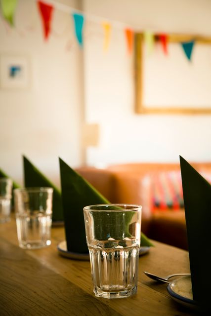 Glass cups and neatly folded green napkins on a wooden dining table inside a cozy room with colorful bunting in the background. Ideal for illustrating party preparations, dining settings, or cozy home interiors.