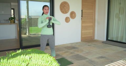 Asian woman working out with kettlebell on sunlit patio in mint-green top