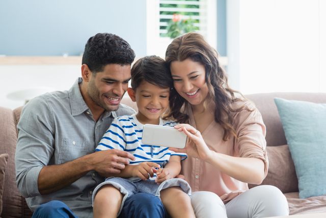 This image shows a happy family of three sitting on a couch, looking at a smartphone together. The parents and their young son are smiling and enjoying their time together. This image can be used for articles or advertisements related to family bonding, parenting tips, technology use in families, or home life. It is also suitable for promoting family-oriented products or services.