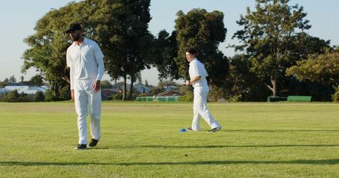 Two Male Cricket Players Practicing on Sunny Grass Field Wearing White Uniforms Near Blue Cone