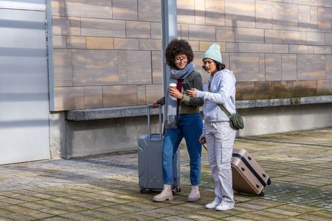 Two women traveling together checking phone while waiting with luggage and coffee