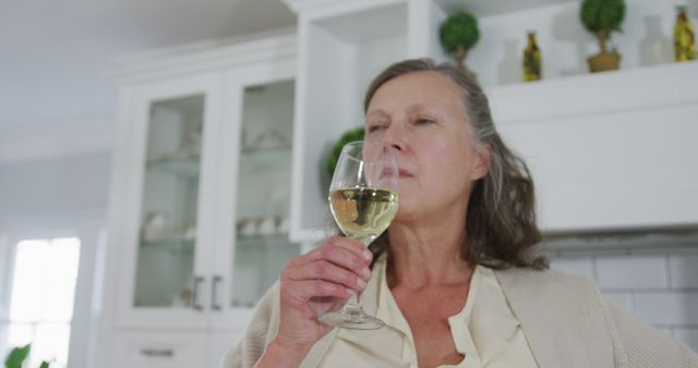 Senior caucasian woman standing in kitchen enjoying drinking a glass of white wine. retirement lifestyle at home.