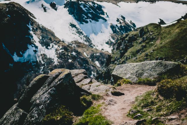 A mountainous trail with large boulders overlooks snow-covered peaks under a clear sky. This picturesque scene is perfect for use in travel brochures, adventure-themed websites, and outdoor activity promotions, capturing the spirit of exploring nature and outdoor adventure.