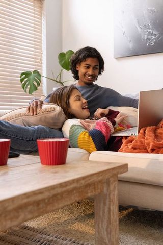 Asian couple relaxing on sofa watching laptop in cozy living room with colorful sweater and red cups