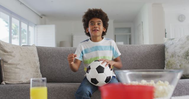 Young boy sitting on a comfortable couch with a soccer ball in hand, watching sports on TV. He appears excited and engaged, showcasing his passion for soccer. Perfect for use in content related to childhood, sports, fan experience, family entertainment, and vibrant home lifestyles.