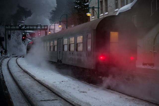 Steam train departs from snowy station with smoke and mist creating atmospheric evening scene. Perfect for themes related to winter travel, transportation history, industrial heritage, adventure and scenic journeys. Ideal for websites, educational materials, and winter travel promotions.