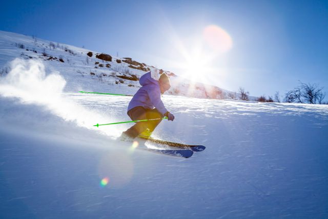 Skier descending snow-covered slope with blue sky and sunshine. Great for winter sport advertisements, travel brochures, adventure magazines, and promotional materials for ski resorts.