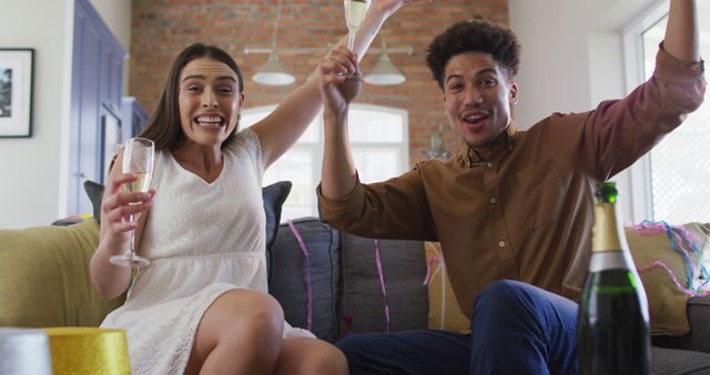 A young couple are celebrating together at home. They are raising champaign glasses while smiling and expressing excitement. This can be used for themes related to celebration, togetherness, relationships, happiness, and special moments.