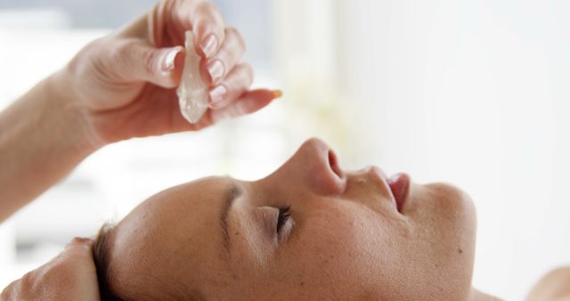 A middle-aged woman is receiving a facial treatment with a clear crystal, with copy space. This wellness practice suggests a spa or therapeutic setting focused on skincare and relaxation.