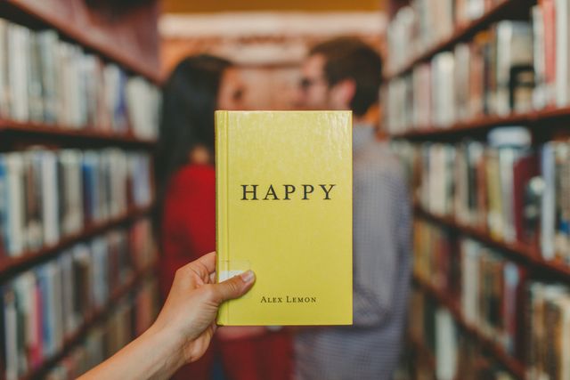 This image shows a person's hand holding a book with the title 'Happy' in a library. The focus on the book contrasts with blurred background featuring shelves filled with books and two people conversing. Great for themes of reading, education, study, book clubs, libraries, or literature-themed content.