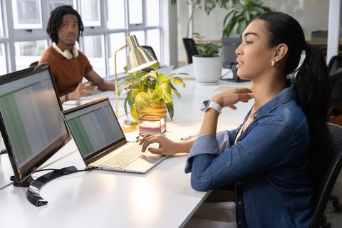 Young professional woman working on laptop with dual monitors in bright open-plan office