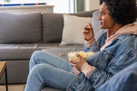 African american woman relaxing on sofa eating popcorn and watching tv in cozy living room