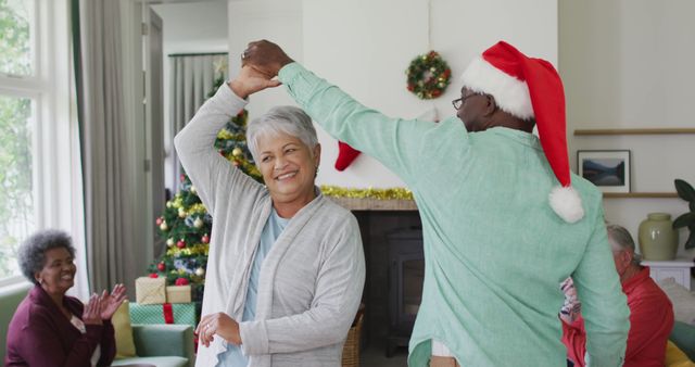 A happy senior couple is dancing together in a festive, decorated living room during a Christmas family gathering. Both are wearing Santa hats, and there are other family members visible in the background, clapping and enjoying the moment. This image is perfect for holiday marketing, greeting cards, or illustrating holiday joy and togetherness in promotional materials.
